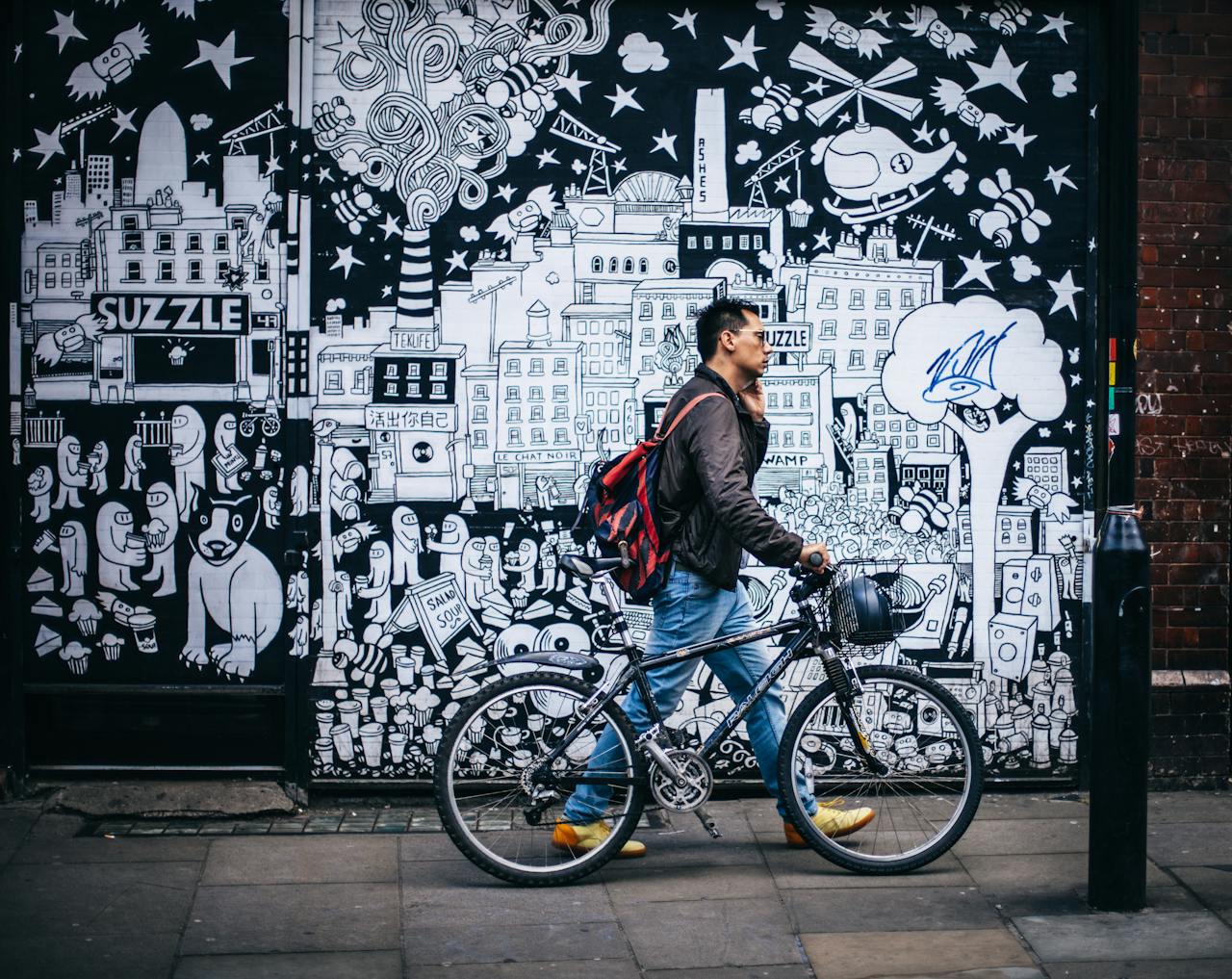 Man walking a bicycle past a detailed urban graffiti mural in a city setting.