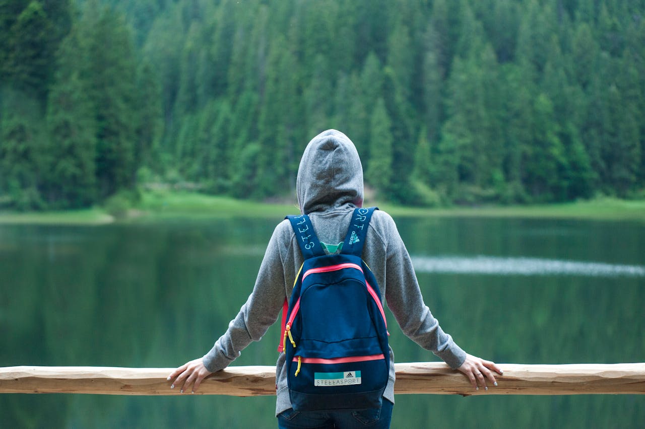 our-story A person in a hoodie stands by a serene lake surrounded by lush greenery, enjoying the peaceful outdoors.